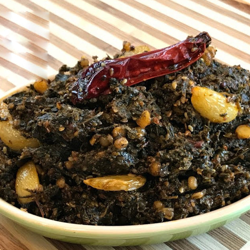A bowl of Akula Gongura Pickle, with visible green and red chili pieces, served on a white plate with a red pickled chili on top.