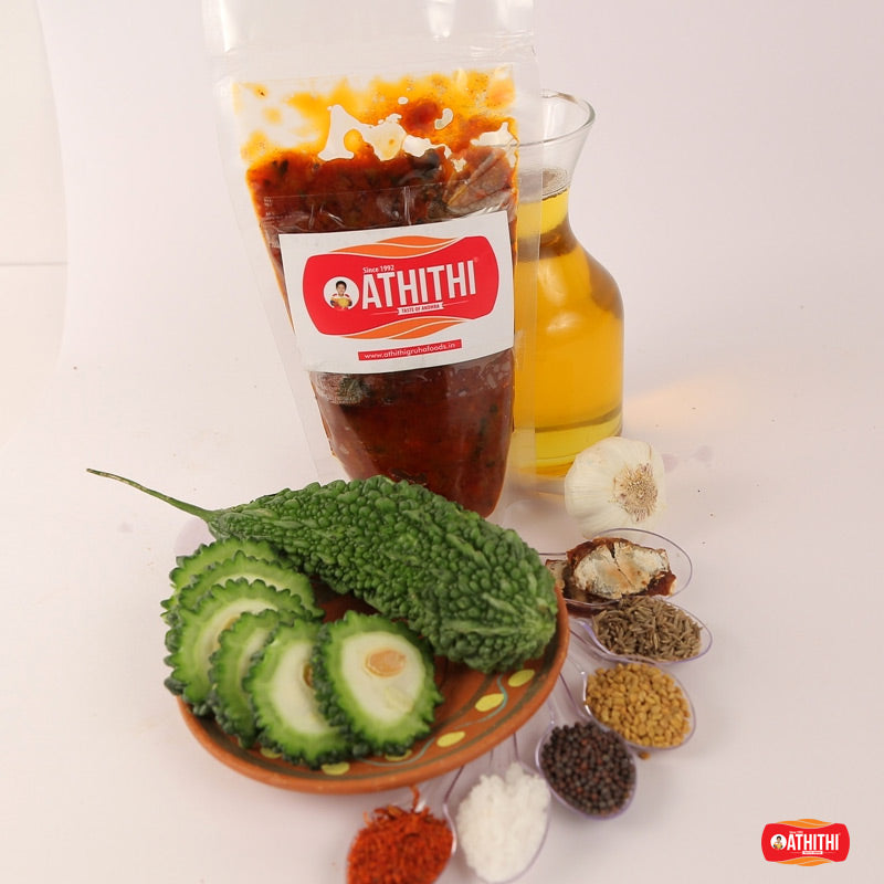 A jar of Kakarakaya Pickle with its label, alongside a bowl of cut cucumbers, spices, and a small oil lamp.