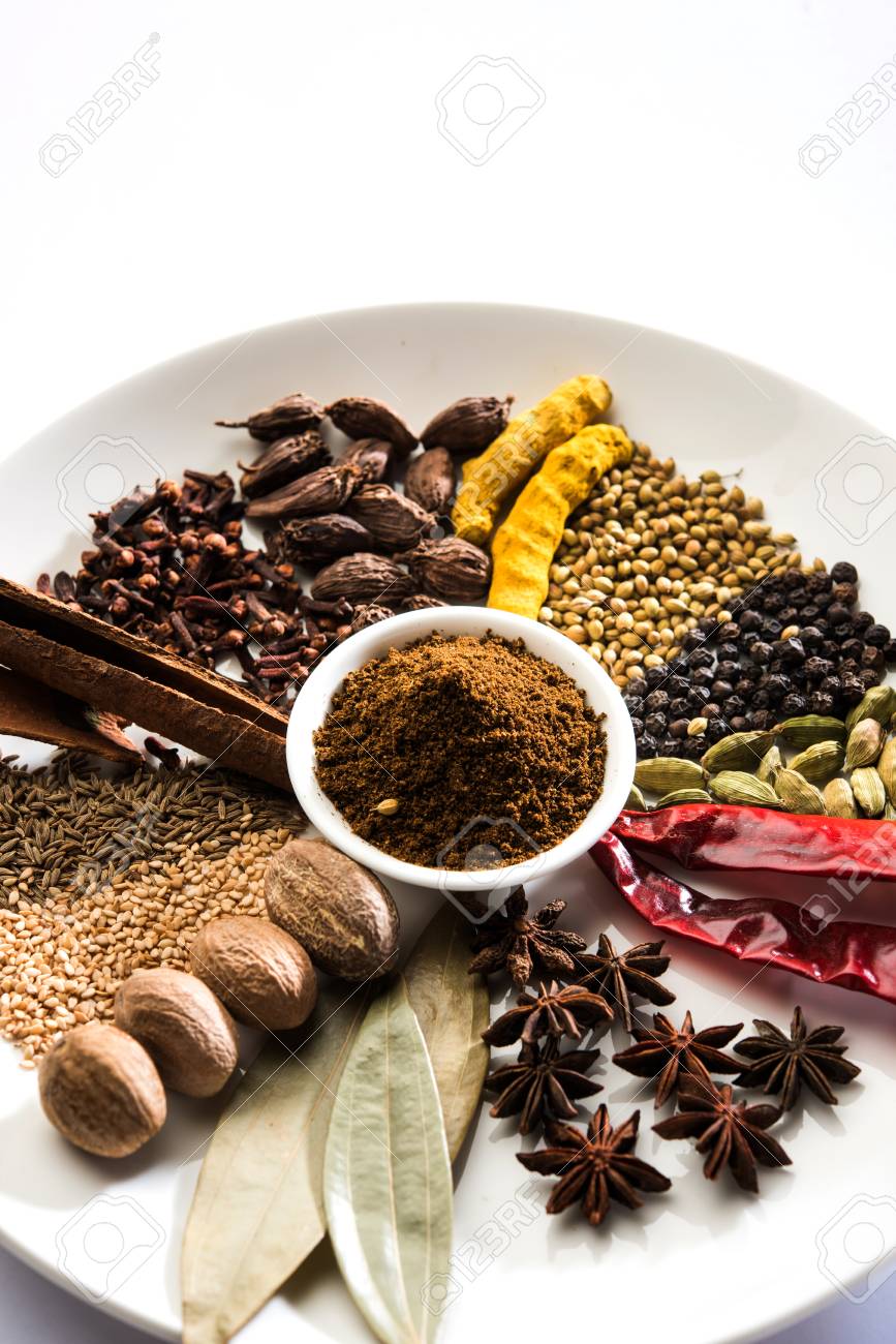 A variety of whole spices and seasonings including cinnamon sticks, cloves, cardamom, cumin, coriander, and whole peppercorns arranged in a bowl on a white plate with a small pile of brown garam masala powder in the center.